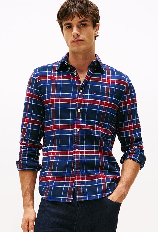 Model wearing a Tommy Hilfiger multicolor plaid button-down shirt with rolled sleeves and denim jeans, standing against a white background.