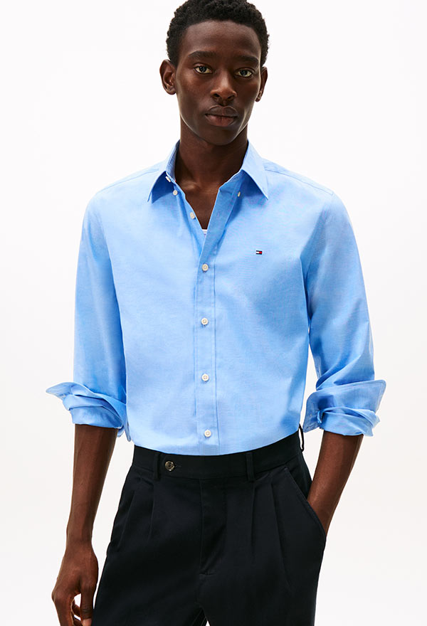 Model wearing a Tommy Hilfiger light blue button-down shirt with short sleeves and dark pants, posed against a white background.