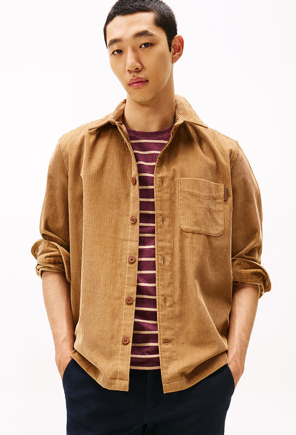 Model wearing a Tommy Hilfiger camel brown corduroy overshirt layered over a striped top, paired with dark trousers, posed against a white background.