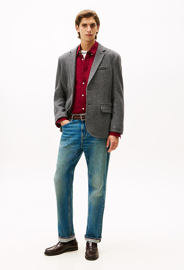 Model wearing a Tommy Hilfiger tailored gray blazer with a red shirt and blue jeans, styled with brown dress shoes, posed against a white background.