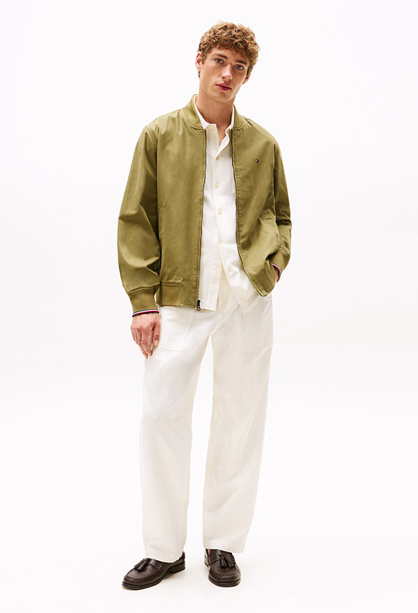 Model wearing a Tommy Hilfiger olive green sherpa zip-up jacket layered over a brown striped top, styled with white pants, standing against a white background.