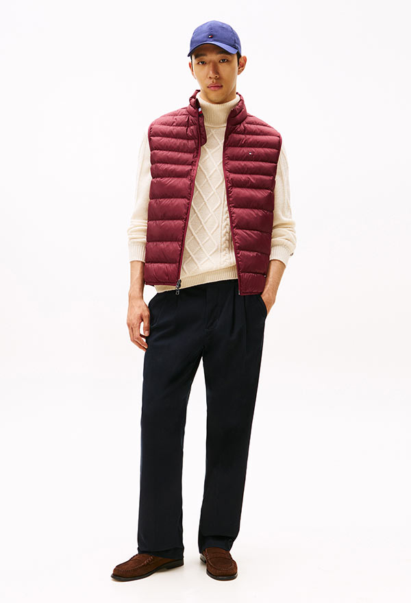 Model wearing a Tommy Hilfiger burgundy quilted vest over a white hoodie, paired with black pants and a blue baseball cap, standing against a white background.