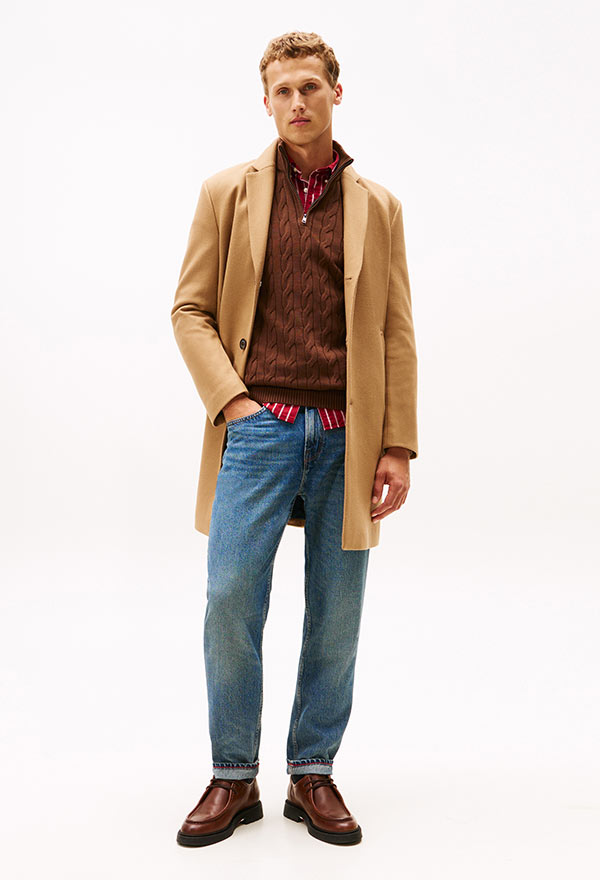 Model wearing a Tommy Hilfiger camel overcoat with a checkered shirt and blue jeans, styled with brown loafers, posed against a white background.