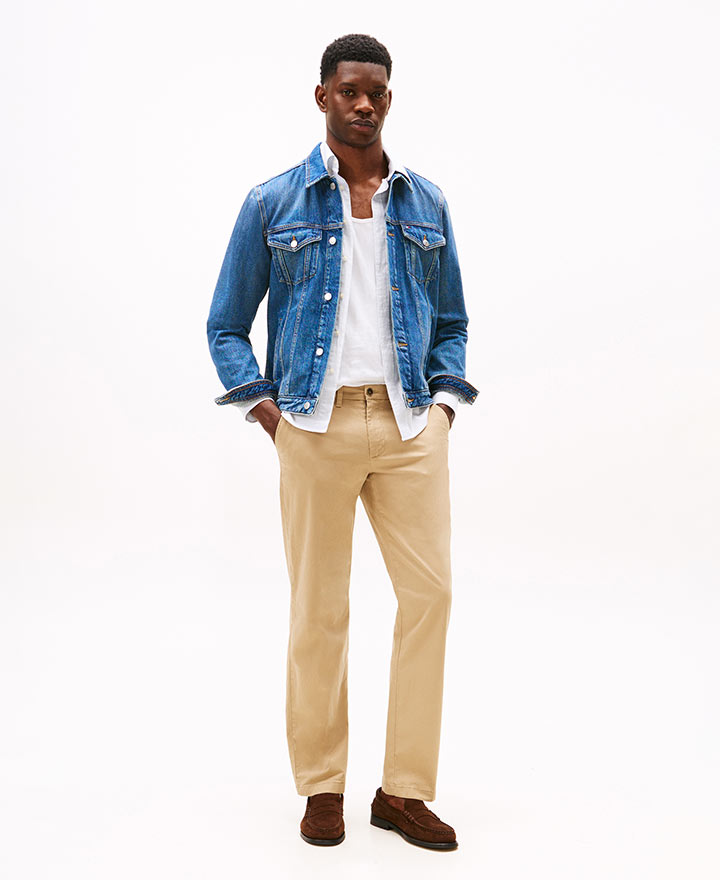 Model wearing a Tommy Hilfiger light blue denim button-down shirt layered over a white graphic tee, paired with khaki pants and brown loafers, standing against a white background.