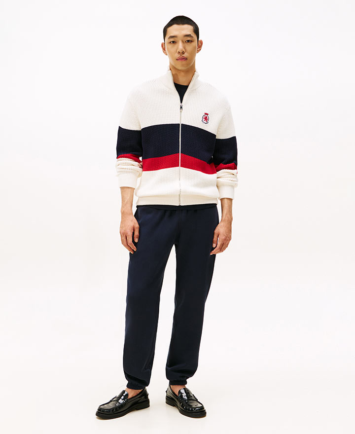 Model wearing a Tommy Hilfiger colorblock pullover sweatshirt in navy, white, and red with the signature logo, paired with navy joggers and black shoes, standing against a white background.