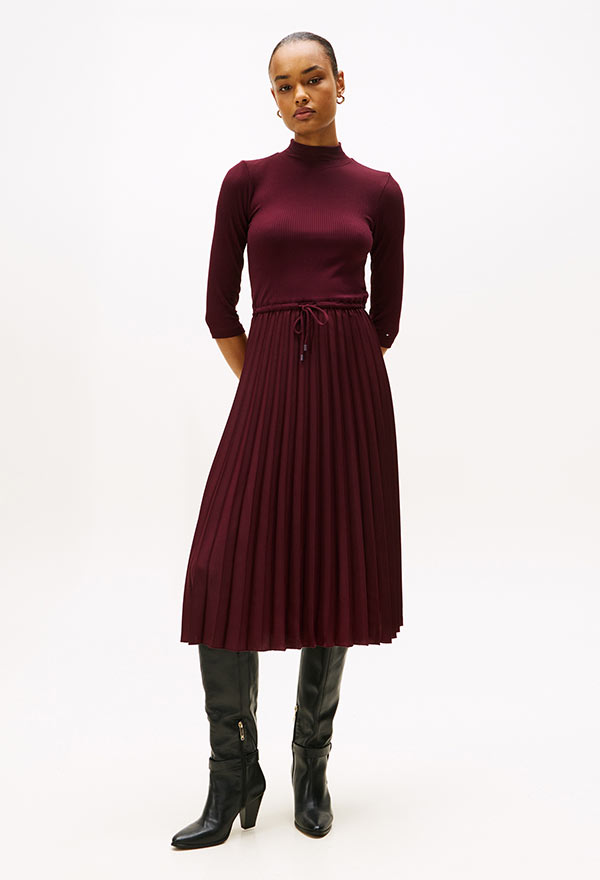 Model wearing a Tommy Hilfiger burgundy long-sleeve pleated midi dress, paired with black boots, standing against a white background.