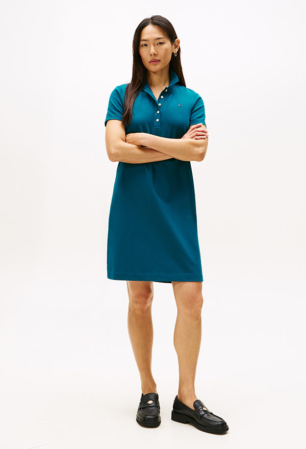 Model wearing a Tommy Hilfiger teal short-sleeve T-shirt dress with logo detailing, styled with black shoes, posed against a white background.