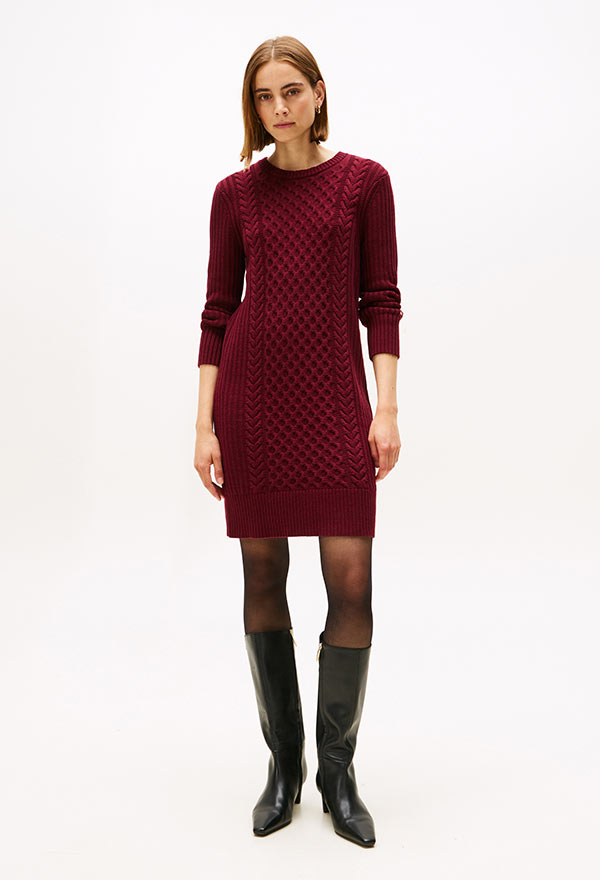 Model wearing a Tommy Hilfiger burgundy knit sweater dress with long sleeves, paired with black knee-high boots, standing against a white background.