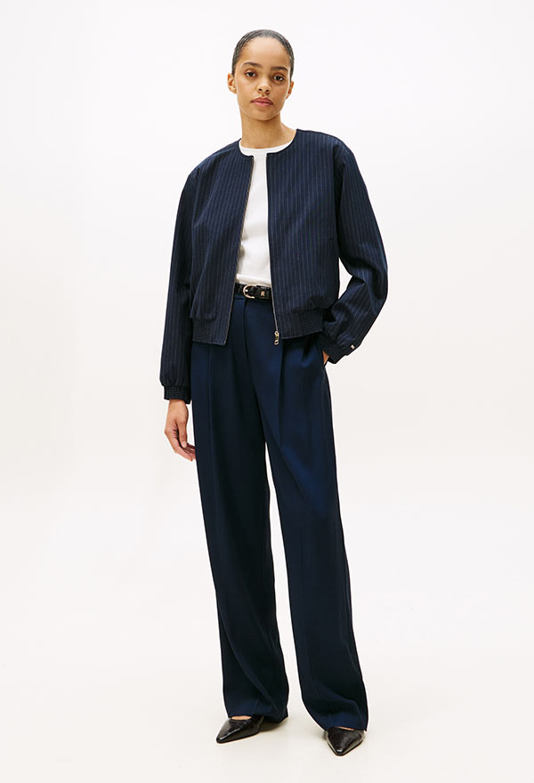 Model wearing a Tommy Hilfiger navy bomber jacket over a white button-down and dark trousers, standing against a white background.