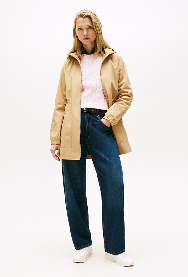 Model wearing a Tommy Hilfiger light tan lightweight jacket over a cream sweater and dark blue jeans with white sneakers, posed against a white background.