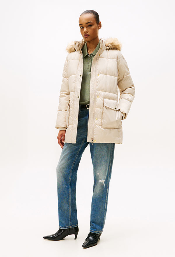 Model wearing a Tommy Hilfiger cream puffer jacket over a striped top, paired with medium-wash jeans and black shoes, standing against a white background.