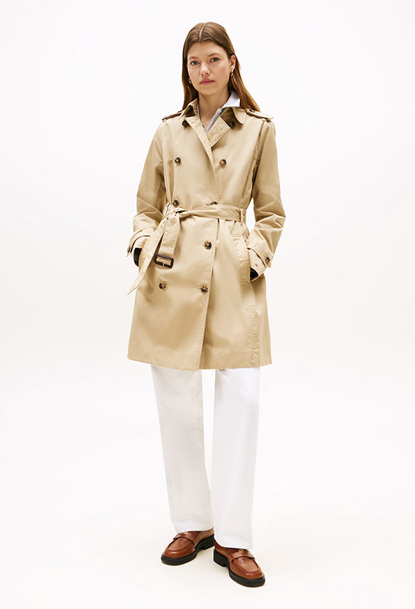 Model wearing a Tommy Hilfiger beige belted trench coat with flap pockets and epaulets, styled with brown loafers, standing against a white background.