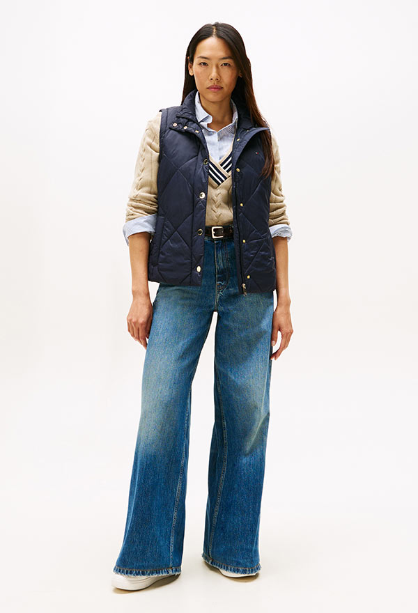 Model wearing a Tommy Hilfiger navy puffer vest layered over a long-sleeve top with wide-leg denim, posed against a white background.