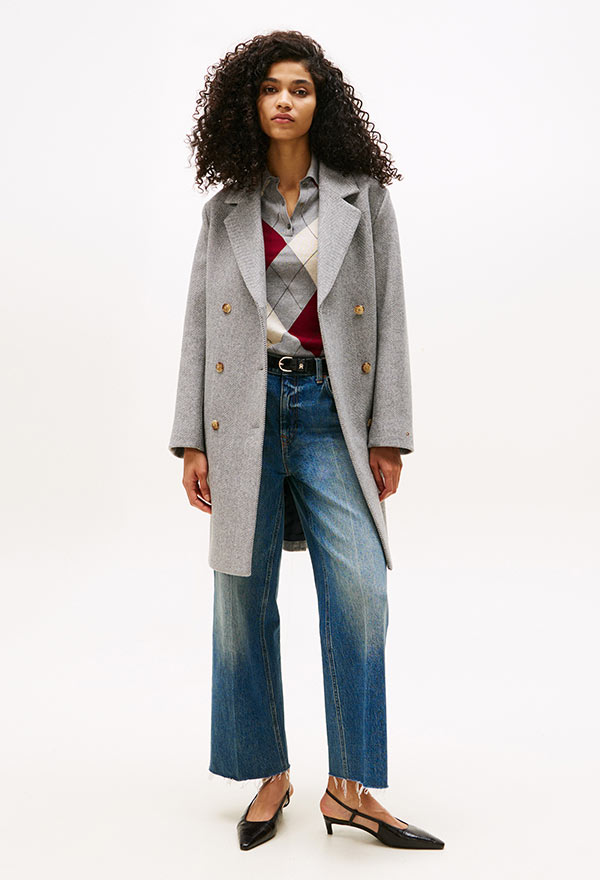 Model wearing a Tommy Hilfiger grey wool coat with a belted waist, styled over a striped shirt and wide-leg jeans, posed against a white background.