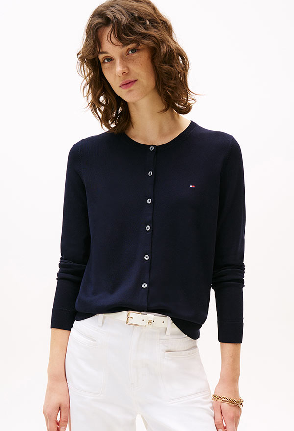 Model wearing a Tommy Hilfiger navy button-up cardigan over a white top, paired with white pants, standing against a white background.