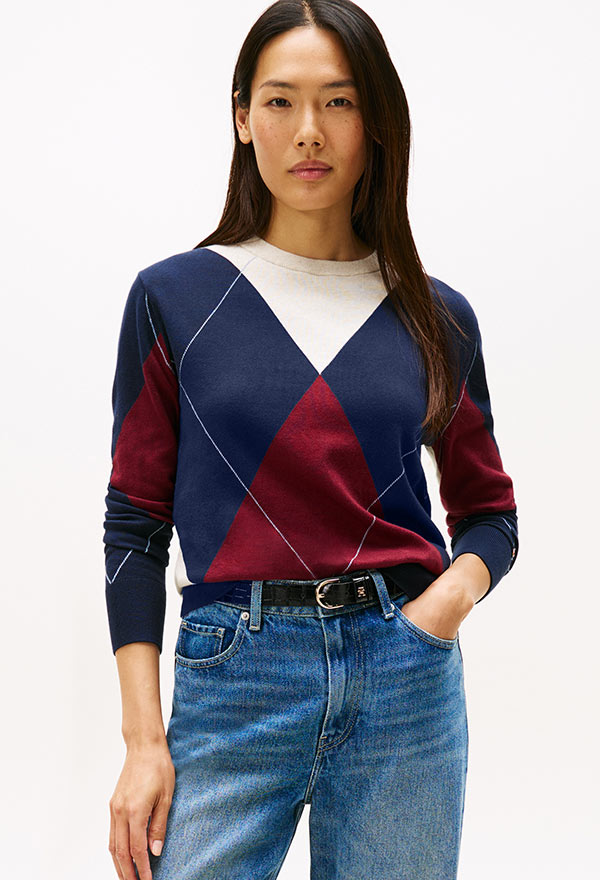 Model wearing a Tommy Hilfiger multicolor V-neck patterned sweater in navy, burgundy, and cream, styled with a denim skirt, posed against a white background.
