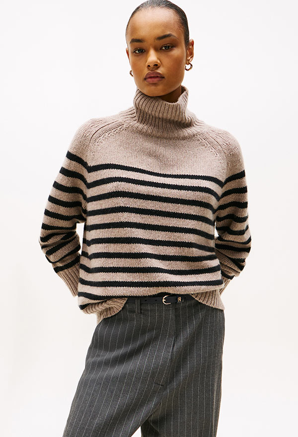 Model wearing a Tommy Hilfiger wool-blend striped sweater in neutral and burgundy tones, styled with a gray skirt, standing against a white background.