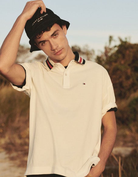 Tommy Hilfiger male model wears white polo layered over blue polo with tan pants and blue hat