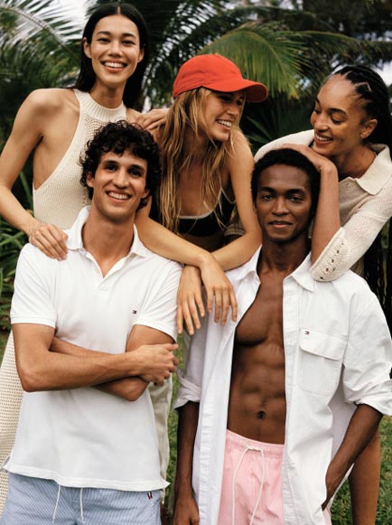 A group of male and female models wear Tommy Hilfiger shirts and shorts on the beach.