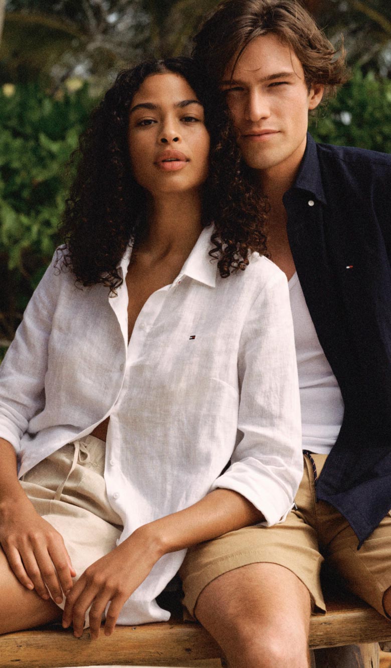 A male and female model wear Tommy Hilfiger linen shirts on the beach.