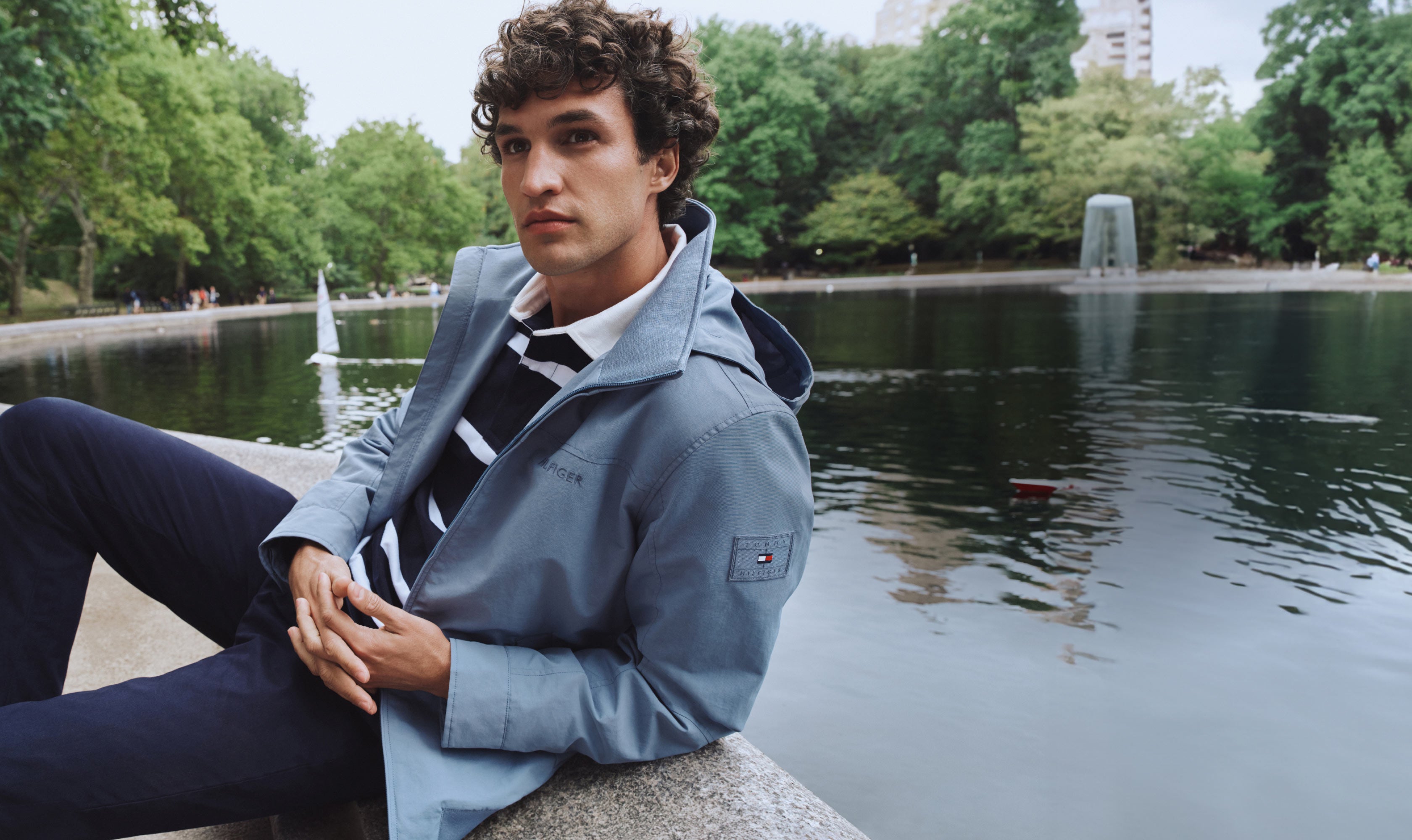 Man wearing Tommy Hilfiger jacket while leaning on pond in the park
