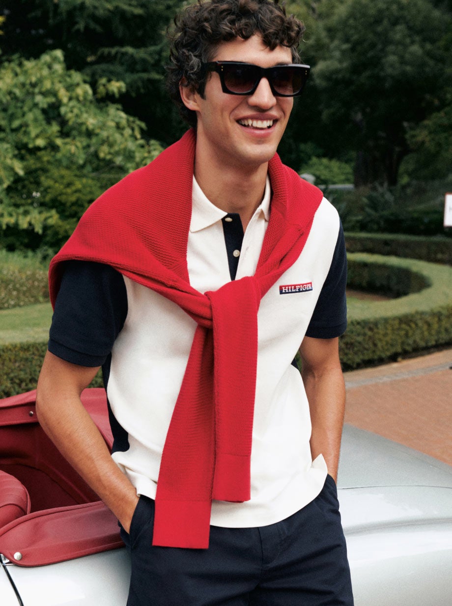 Man leaning on a car with sunglasses on wearing Tommy Hilfiger polo and a sweater over his shoulders