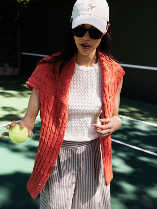 Woman on the tennis court wearing Tommy Hilfiger Hat and tshirt with a sweater over her shoulders