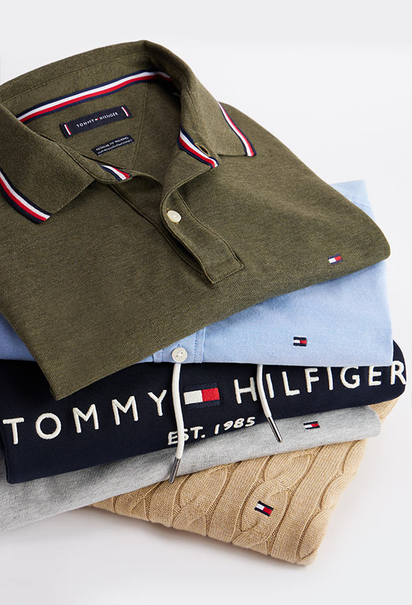 Stack of folded Tommy Hilfiger men's shirts, including an olive green polo, light blue button-down, navy logo tee, gray sweatshirt, and beige cable-knit sweater, showcasing signature flag branding
