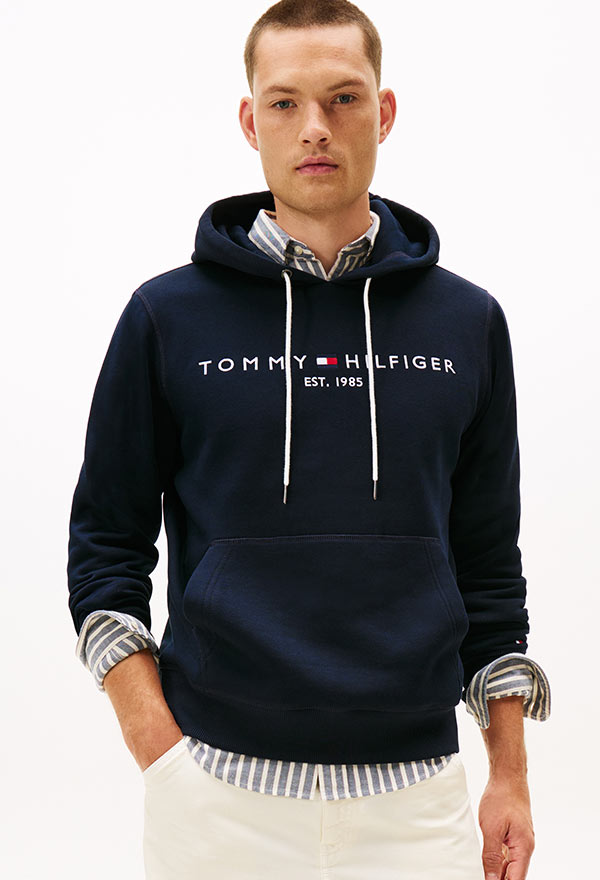 Model wearing a Tommy Hilfiger navy pullover hoodie with bold logo across the chest, paired with white pants, standing against a white background.