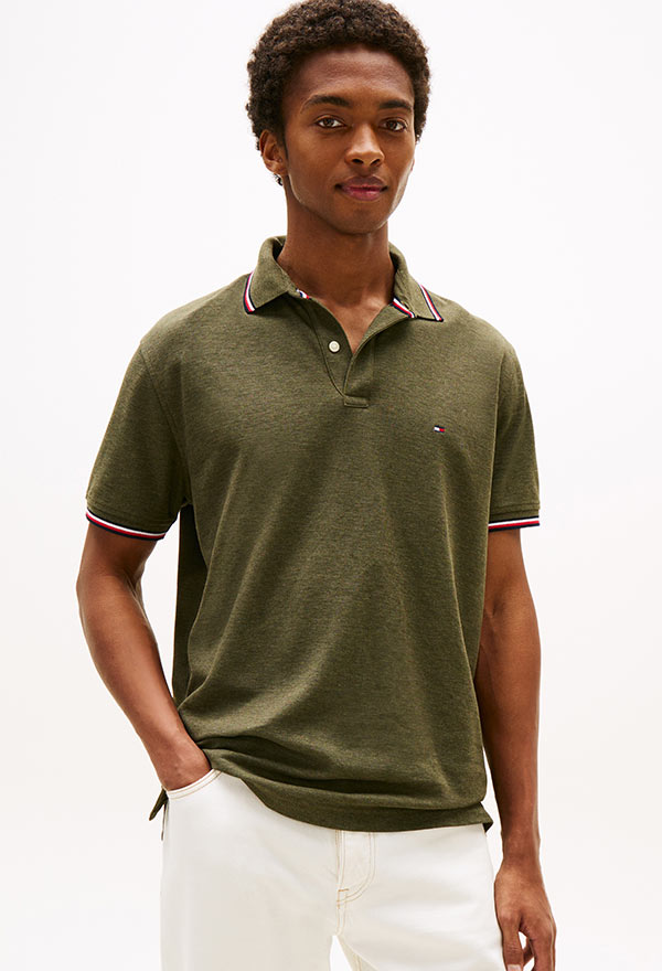 Man wearing a beige cable-knit sweater layered over a striped button-down shirt, paired with white pants, standing against a white background.