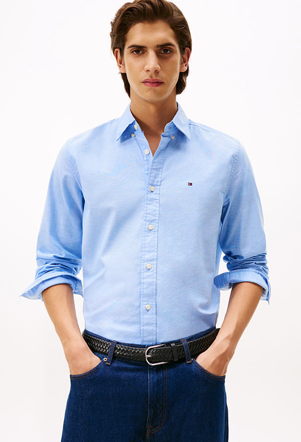 Model wearing a Tommy Hilfiger light blue long-sleeve button-down shirt with embroidered logo, paired with dark jeans, standing against a white background.