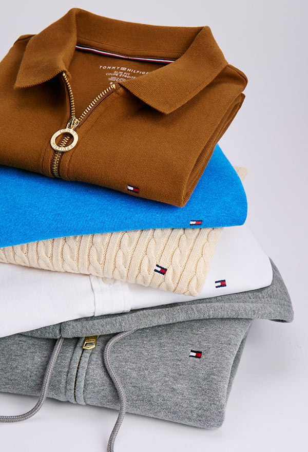 Stack of folded Tommy Hilfiger women’s tops including a camel polo, blue sweater, grey knit, and navy piece, each featuring distinct textures and logo accents.