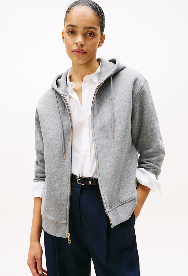Model wearing a Tommy Hilfiger light grey zip-up hoodie layered over a white T-shirt and paired with blue jeans, posed against a white background.