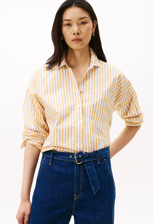 Model wearing a Tommy Hilfiger beige collared shirt with rolled sleeves, styled with a high-waisted denim skirt, posed against a white background.