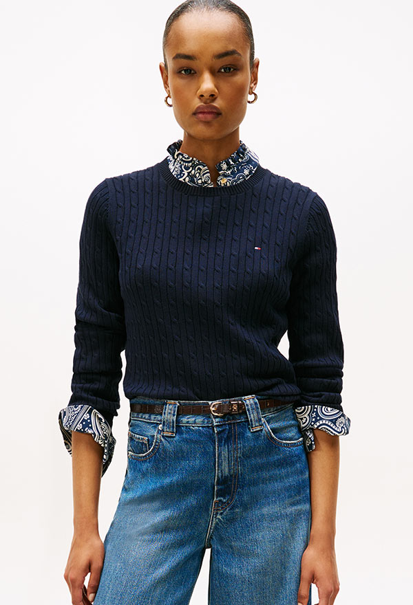 Model wearing a Tommy Hilfiger navy crewneck sweater with rolled sleeves and light blue jeans, standing against a white background.