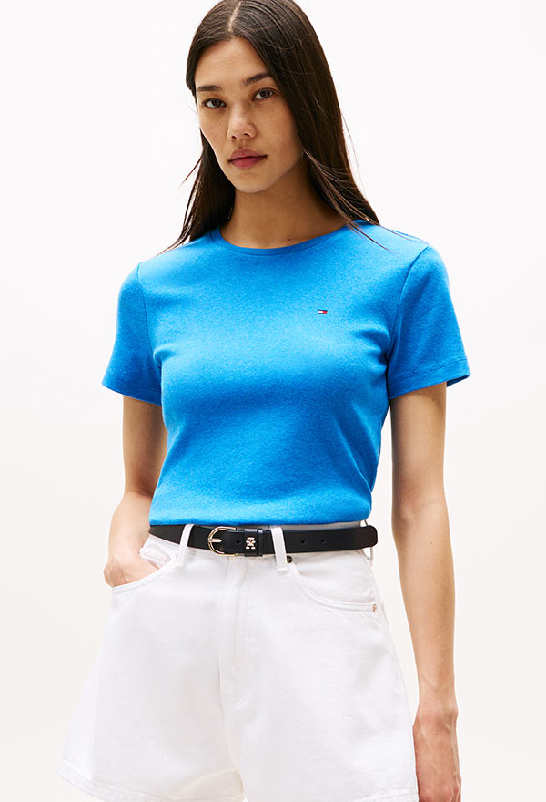 Model wearing a Tommy Hilfiger bright blue crewneck T-shirt tucked into high-rise white pants, standing against a white background.