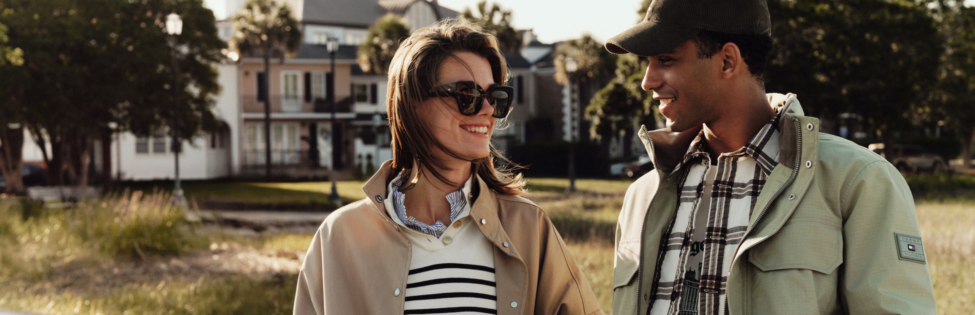 Man and woman in neutral-toned outerwear, styled with striped tops and scarves, walking outdoors near modern houses.