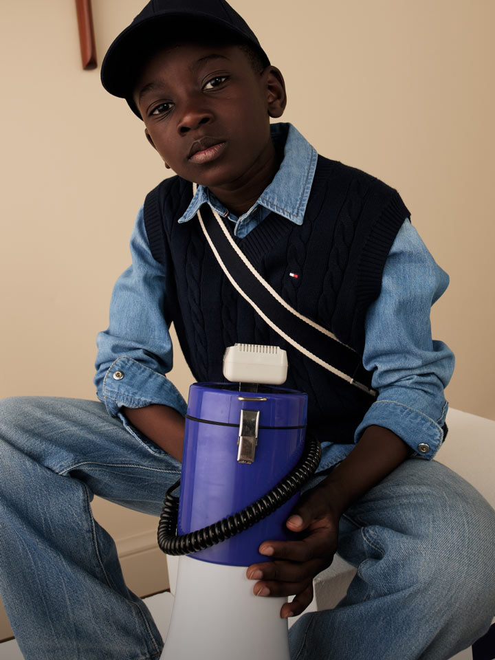 Young boy wearing a yellow quilted vest over a blue striped button-down shirt with light-wash jeans and white sneakers, styled in a Tommy Hilfiger kids look, standing in a playroom with vintage decor.