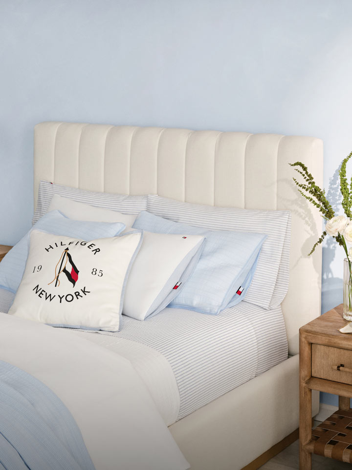 Tommy Hilfiger bedding styled in a serene bedroom with a cream upholstered bed, light blue and white striped sheets, and embroidered pillows—including a white New York-themed accent pillow—paired with natural wood nightstands and woven pendant lighting.