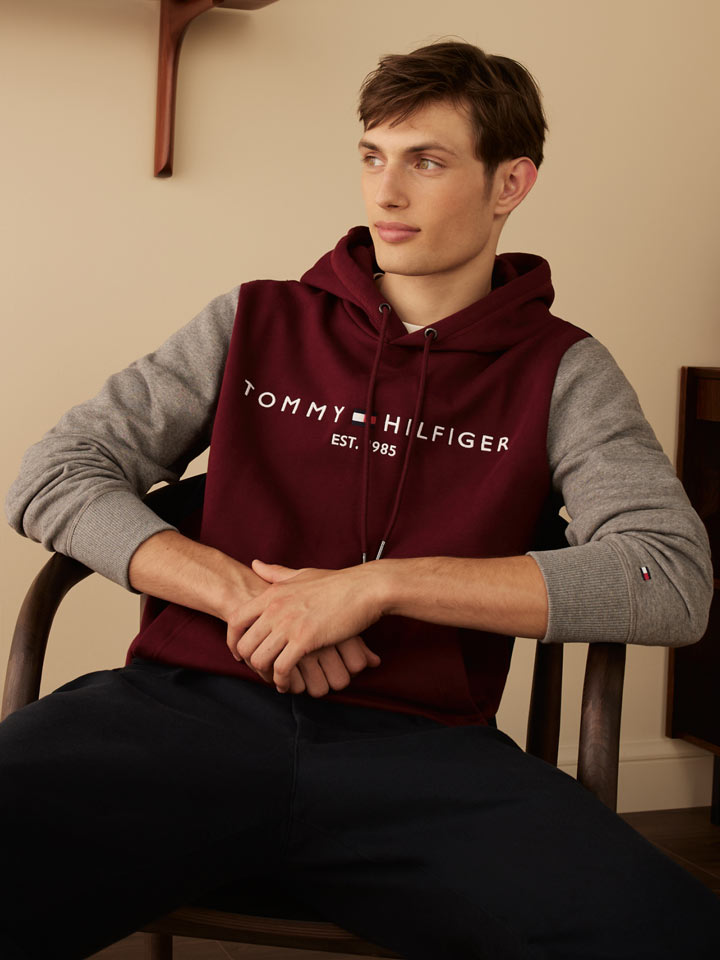 Teen boy wearing a Tommy Hilfiger colorblock hoodie in burgundy and gray, paired with navy pants, seated in a wooden chair in a minimalist room with modern shelving and decor