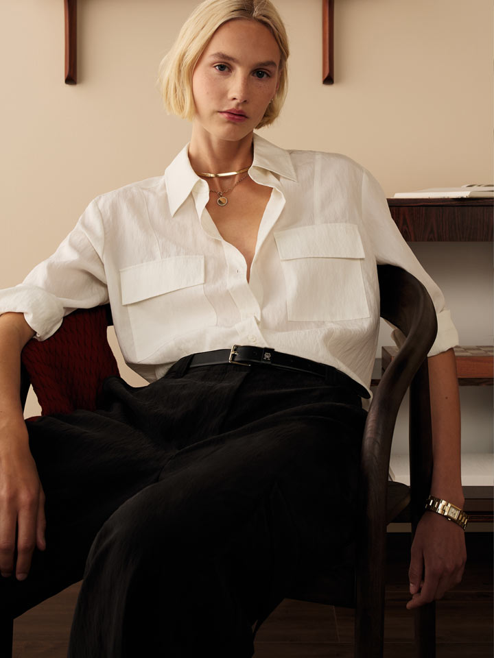 Female model in a relaxed Tommy Hilfiger look, wearing a white button-down shirt with oversized pockets and black wide-leg trousers, seated in a mid-century wooden chair