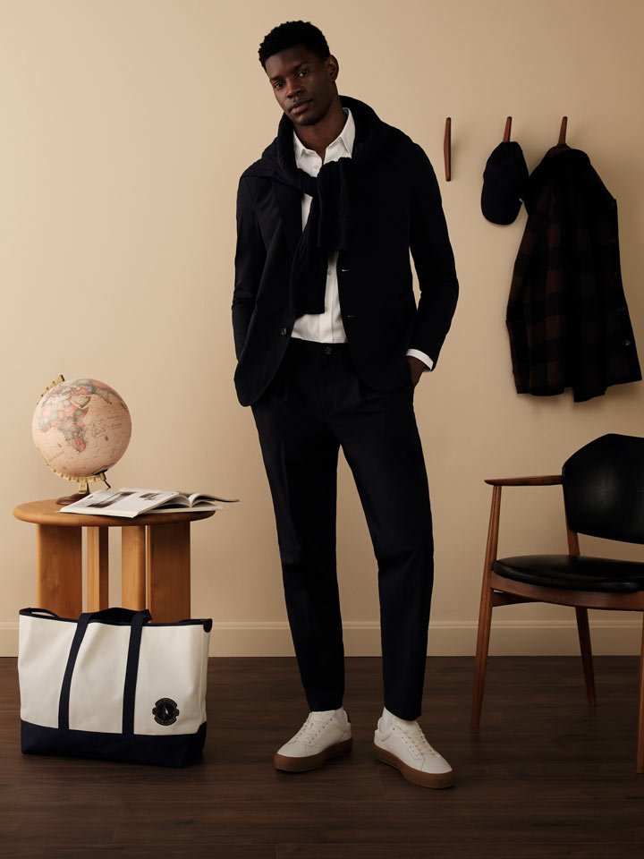 Male model dressed in a refined Tommy Hilfiger outfit, featuring a navy jacket and trousers with a white shirt and slip-on shoes, posed beside a tote bag and globe in a modern, neutral-toned room.