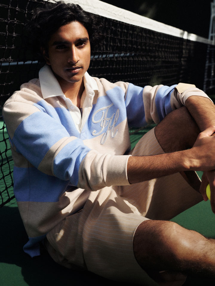 Male model dressed in a refined Tommy Hilfiger outfit, featuring a navy jacket and trousers with a white shirt and slip-on shoes, posed beside a tote bag and globe in a modern, neutral-toned room.