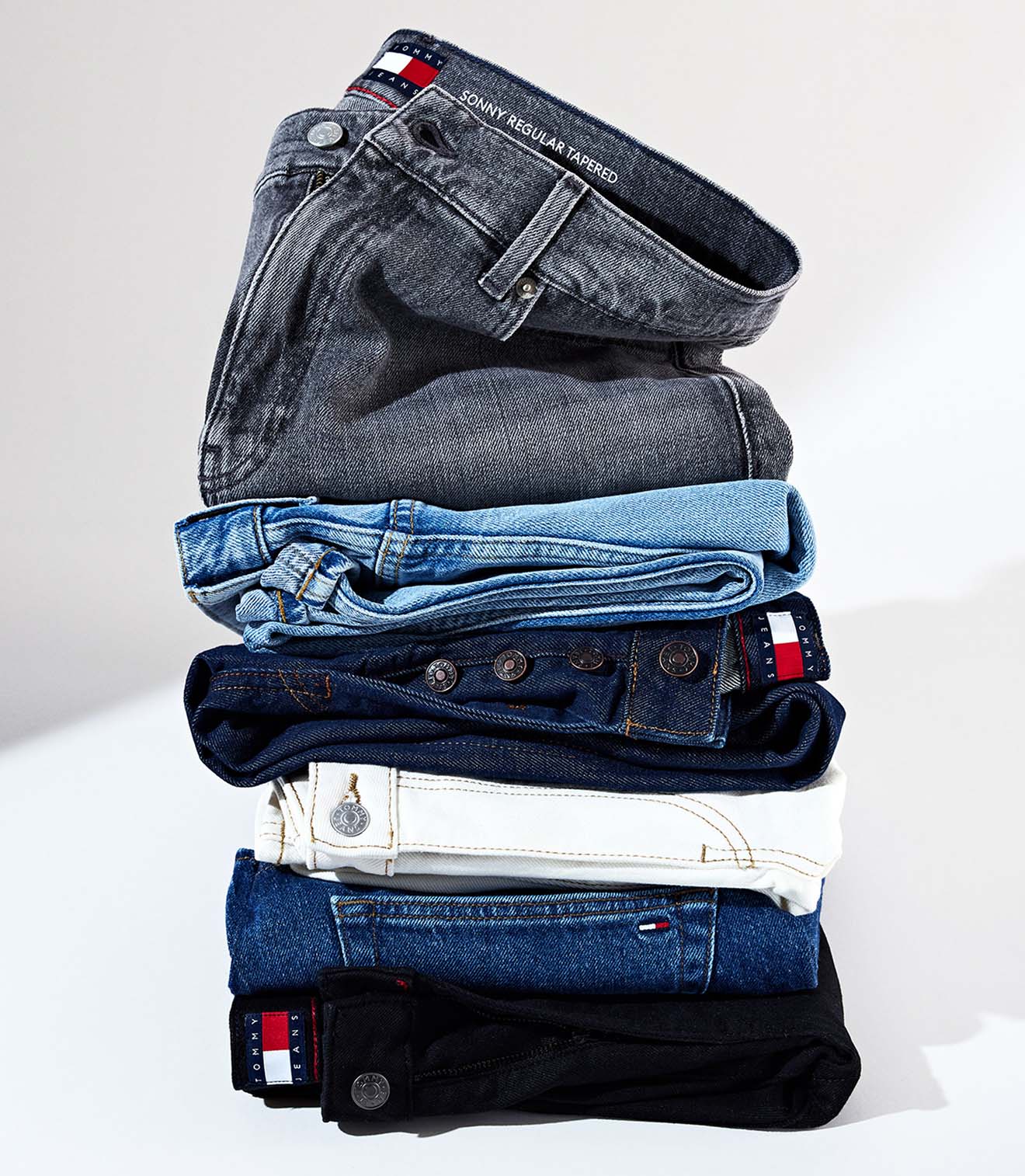 Stack of folded Tommy Jeans denim in assorted washes, including black, indigo, and light blue, each featuring the signature logo tag.