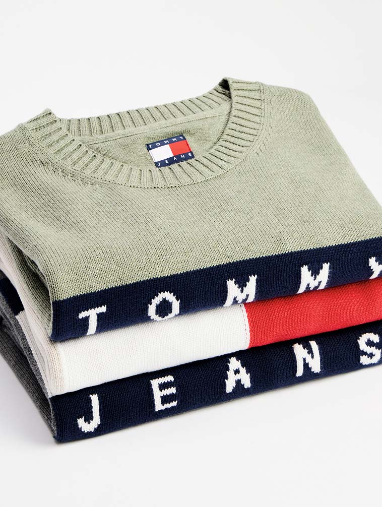Stacked Tommy Jeans crewneck sweaters in soft green and navy, showcasing bold logo lettering and the signature flag patch on the collar.