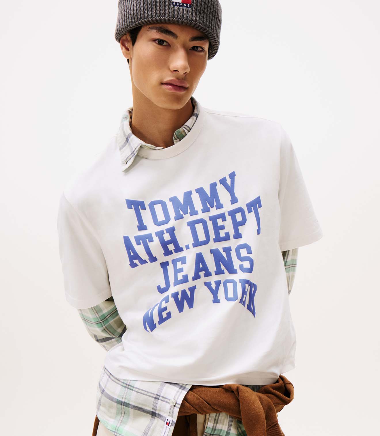Male model wearing a white Tommy Jeans long-sleeve graphic T-shirt with bold blue text, styled with a plaid shirt around the waist and a beanie.