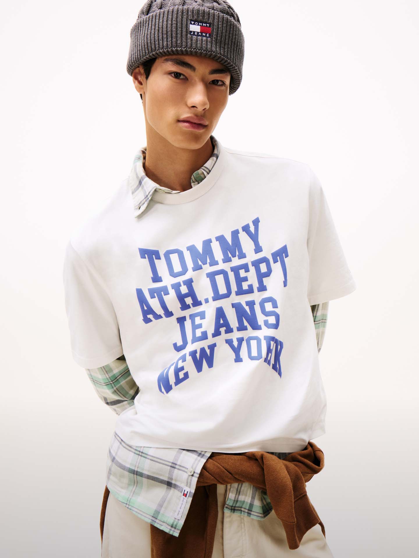 Male model wearing a white Tommy Jeans T-shirt with bold blue collegiate text reading 'Tommy Hilfiger Jeans New York', styled with a flannel shirt tied at the waist and a beanie.