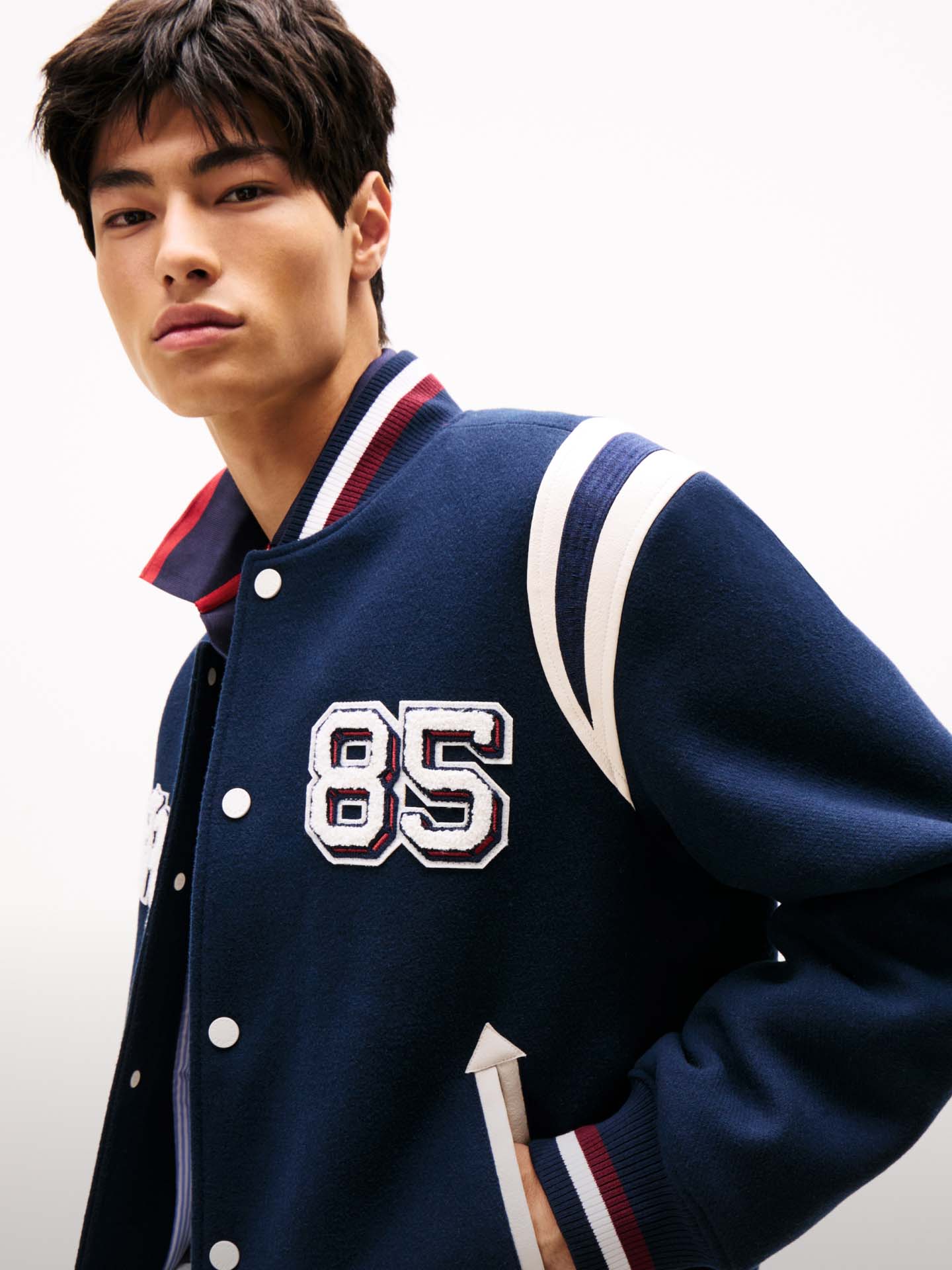 Male model dressed in a gray Tommy Jeans denim jacket and jeans, styled with a red hoodie, logo tee, and baseball cap, seated with a casual pose.
