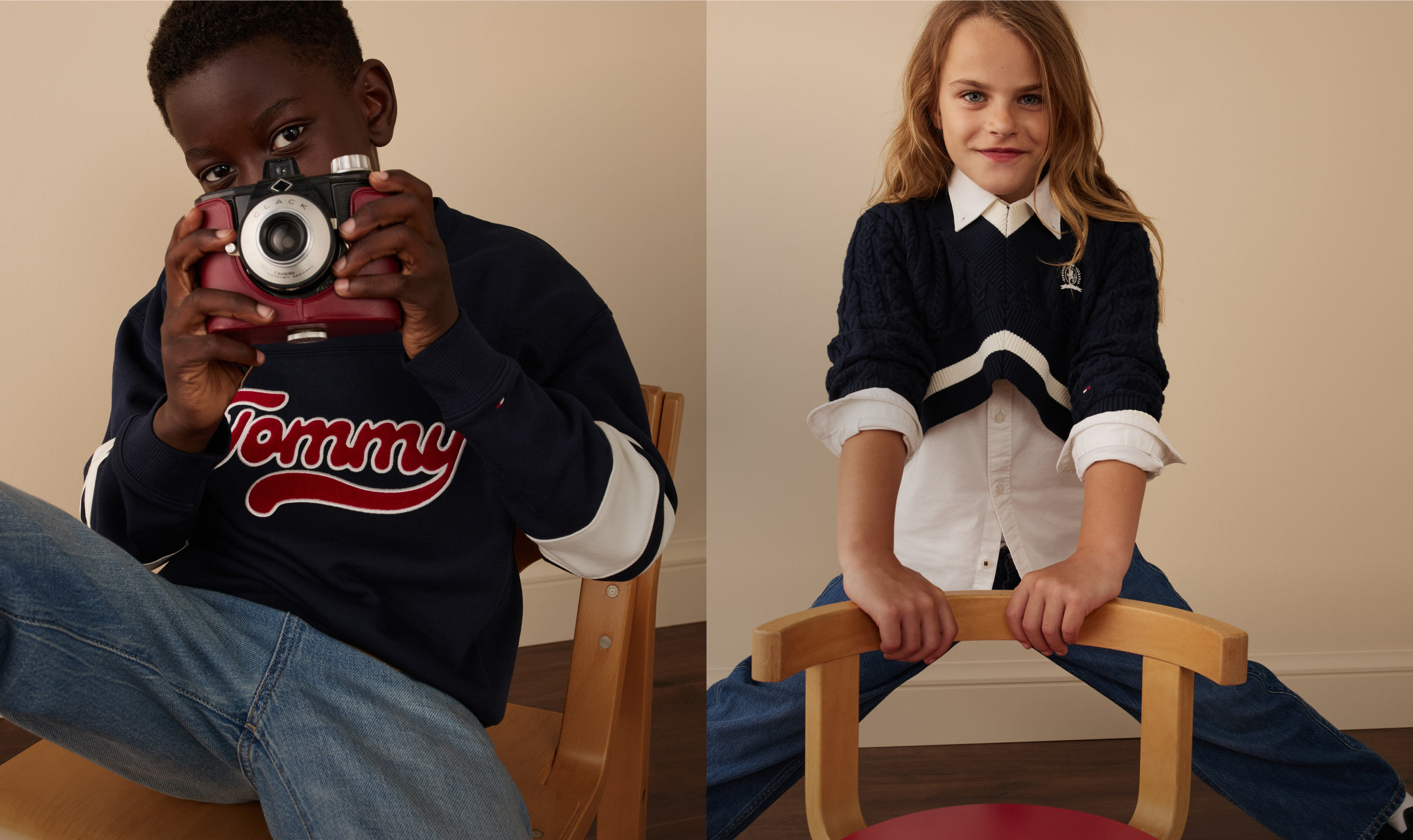 Kids in Tommy Hilfiger back-to-school outfits—one in a logo sweatshirt and jeans holding a camera, the other in a preppy navy sweater with a white collar—sitting indoors.