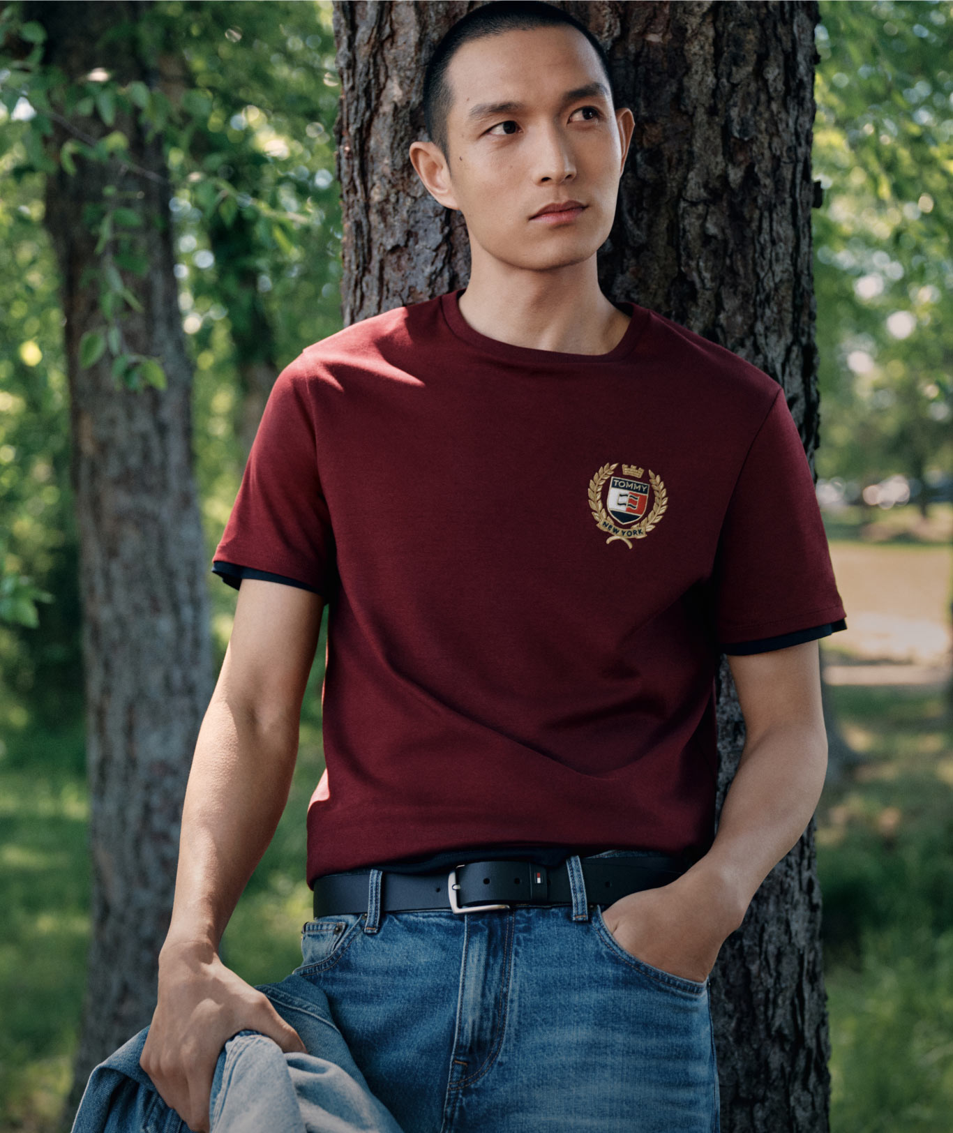 Male model wearing a burgundy Tommy Hilfiger graphic T-shirt and denim jeans, standing outdoors in a forested setting.